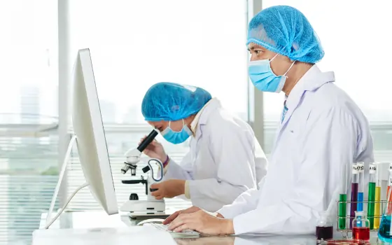 Two lab technicians in protective gear working at computer stations in a laboratory.