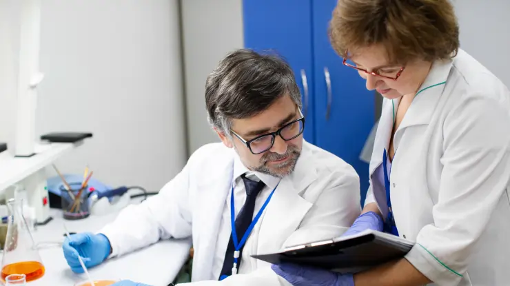 Two pharmaceutical researchers in lab coats reviewing a research document beside lab glassware.