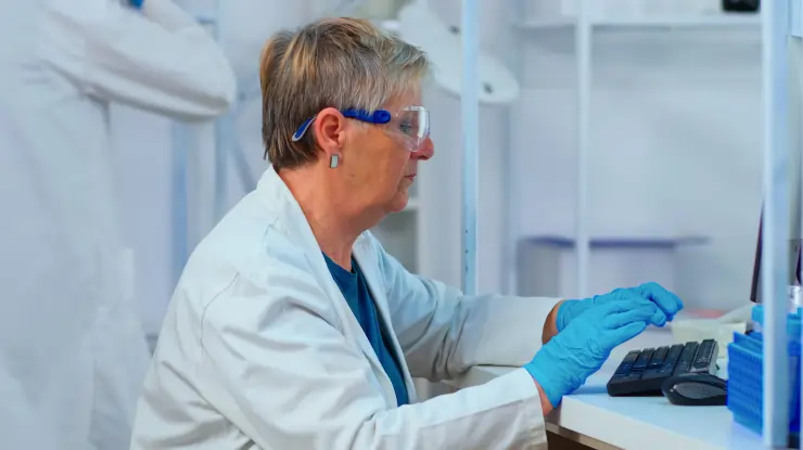 Female laboratory scientist in safety goggles and a lab coat working at a computer for data analysis or research.