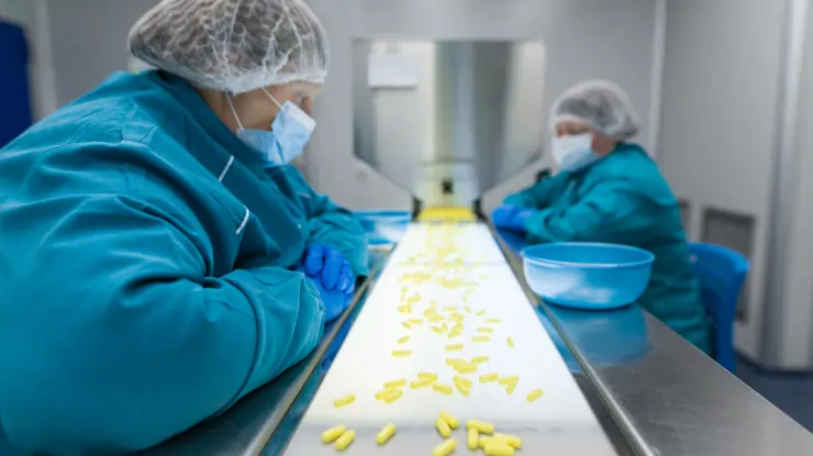 Workers monitoring yellow pills on a conveyor belt in a pharmaceutical manufacturing cleanroom.