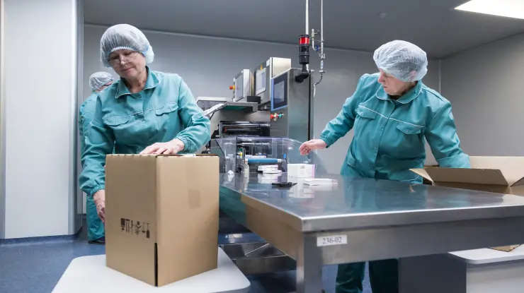 Cleanroom staff in teal uniforms performing packaging and boxing operations for finished     pharmaceutical products.