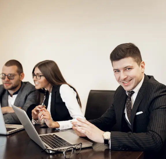 Business professional in a suit collaborating with clients at a desk, smiling while working on a laptop.