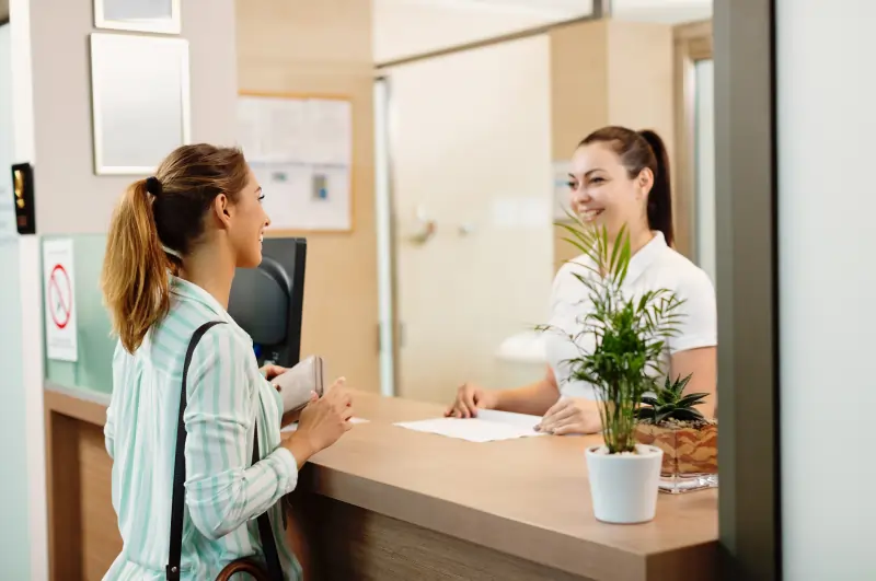 Female patient talking with receptionist at hospital front desk, demonstrating efficient hospital management and patient-friendly workflow with Script All DNA HMS