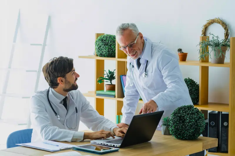 Two male doctors smiling and discussing patient data on a PC screen in hospital, highlighting key features of Script All DNA’s Hospital Management System