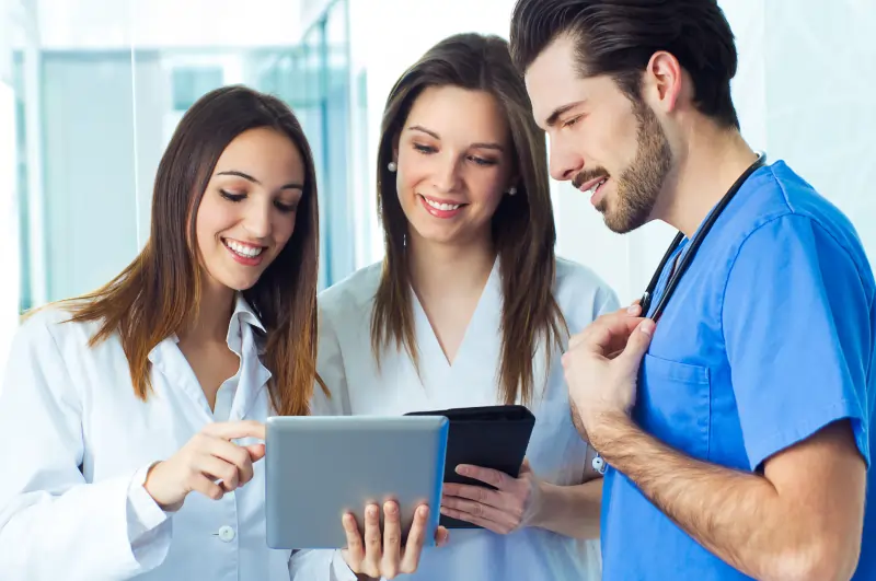 Three young doctors standing and smiling while discussing patient data on a tablet, showcasing collaborative hospital management with Script All DNA HMS