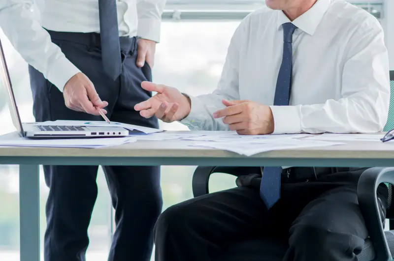 Two business professionals reviewing documents and pointing at data on a laptop screen during an office meeting.