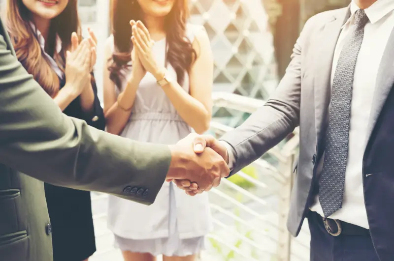 Two male executives shaking hands outdoors while colleagues in the background applaud.