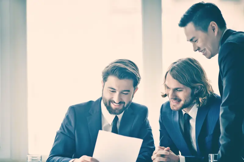 Senior IT professionals collaborating on a laptop in a control room with large digital screens.