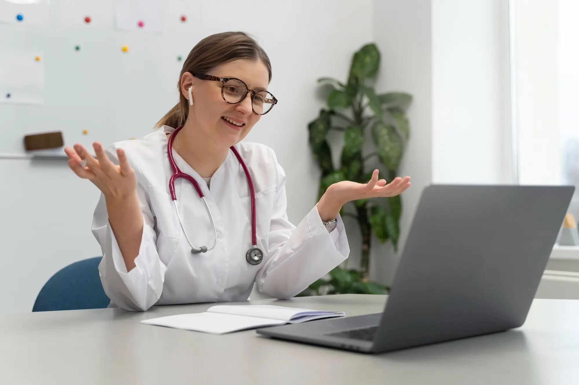Female doctor raising her hands during a video consultation on a laptop while sitting at a desk.